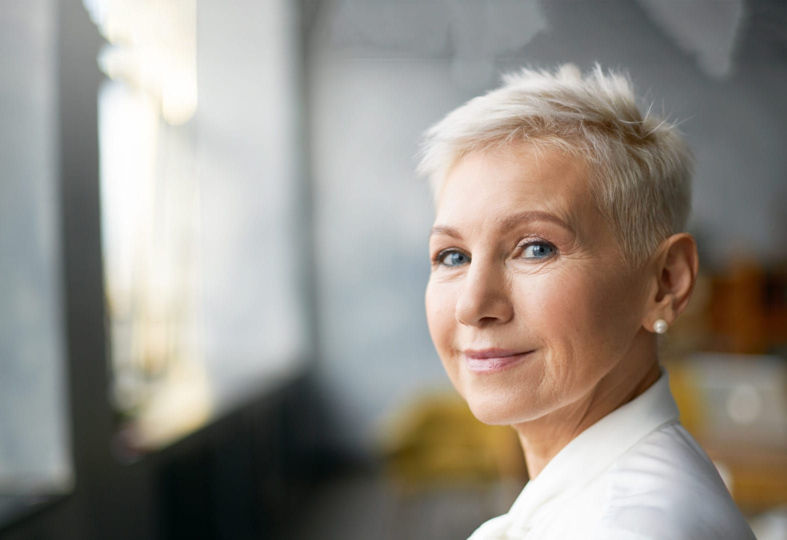 Close Up Portrait Of Attractive Mature Caucasian Woman With Blonde Pixie Haircut And Wrinkles Looking At Camera With Confident Smile While Working In Her Office. Business, Work, Job And Profession